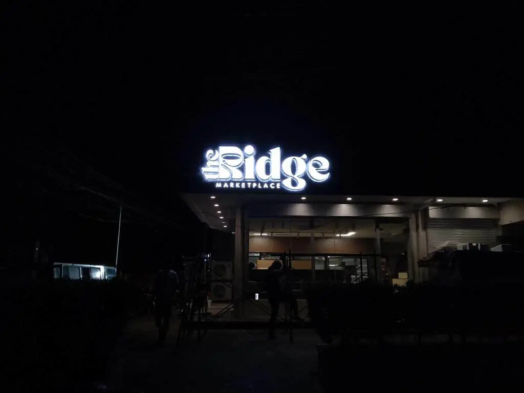 Metallic outdoor signage for The Ridge Marketplace at night in BGC, with people near entrance by M&G Global Ads.