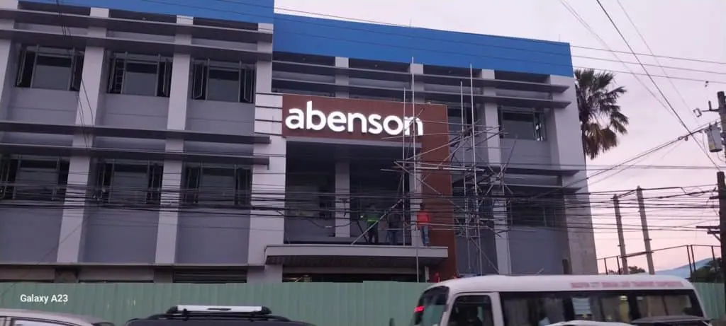Illuminated acrylic abenson signage on a three-story building in Quezon City by M&G Global Ads, scaffolding and cars visible.