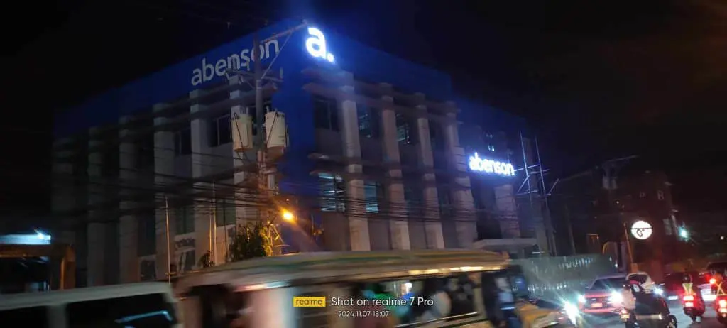 Abenson LED outdoor signage lighting up at night in Quezon City, with busy street traffic and wires by M&G Global Ads.