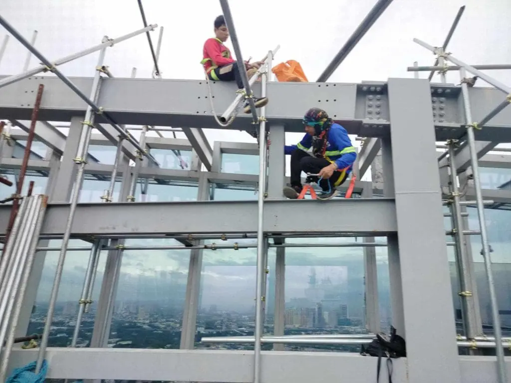 Workers install outdoor billboard signage on a steel frame above Makati skyline, with cloudy sky in background, by M&G Global Ads.