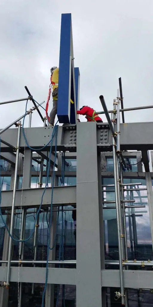 Worker in safety gear installs blue acrylic building signage on scaffolding in Makati, cloudy sky, by M&G Global Ads.