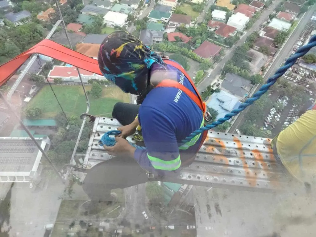 Worker cleans building window on suspended platform beside metallic signage in Ortigas, high above ground level by M&G Global Ads.