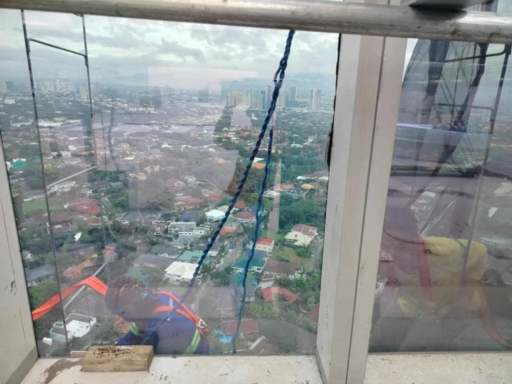 A window cleaner outside a high-rise in Ortigas, with billboard signage and cityscape below, by M&G Global Ads.