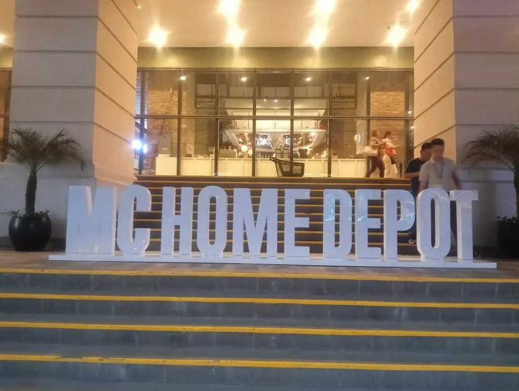 White acrylic MC HOME DEPOT signage installation outside building entrance in Mandaluyong, with plants and people nearby by M&G Global Ads.