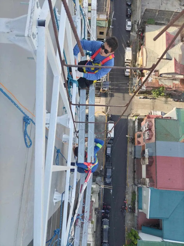 Workers in harnesses install billboard signage on tall metal frame above city street in Makati, buildings below, by M&G Global Ads.