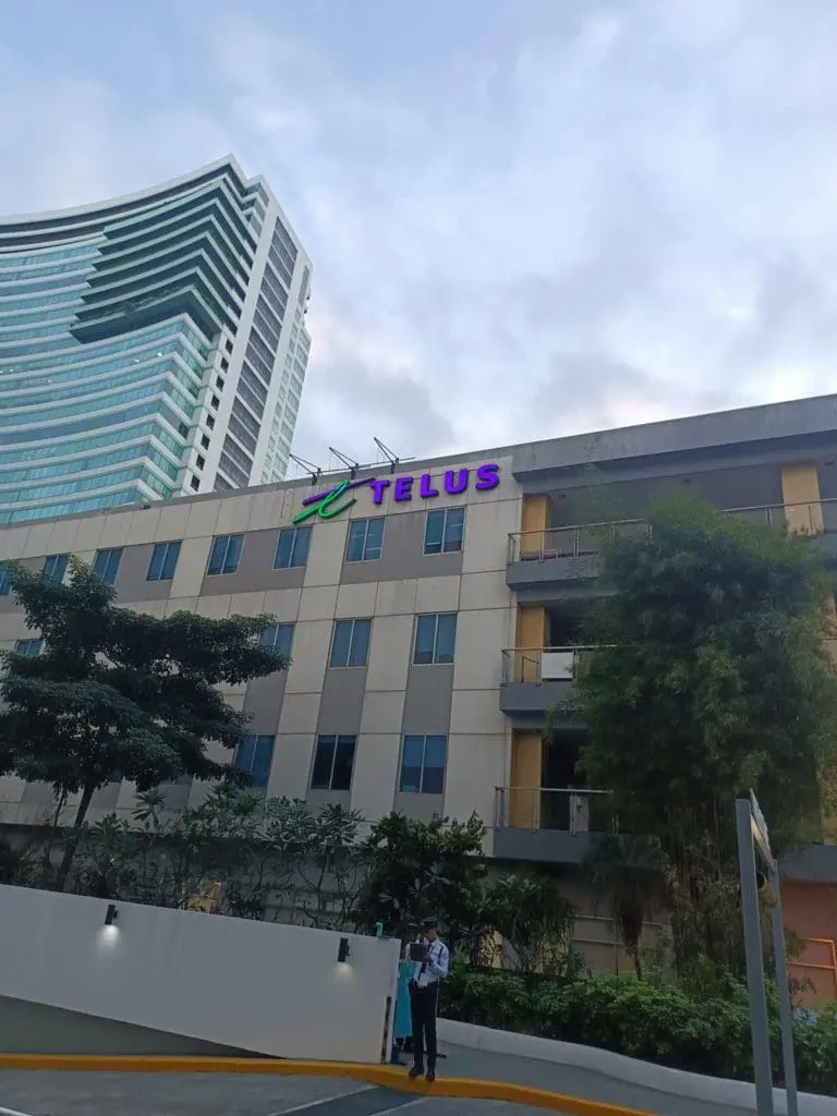 TELUS outdoor building signage in purple and green in Makati, security guard at entrance, glass skyscraper—by M&G Global Ads.