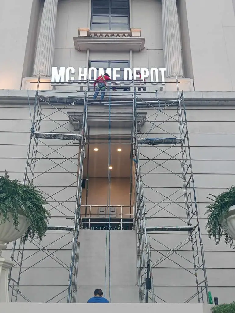 MC Home Depot stainless steel signage installation on building exterior in Pasig, with workers and plants, by M&G Global Ads.