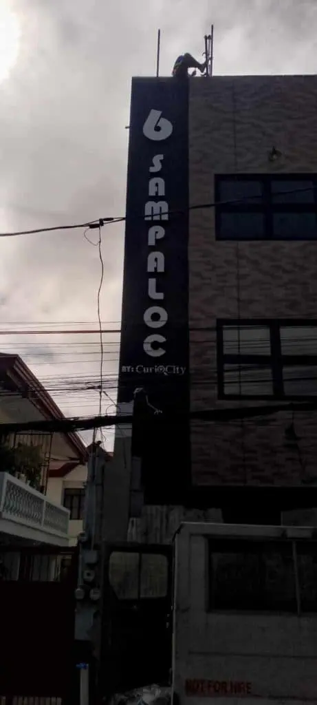 Acrylic building signage for CurioCity, 6 SAMPALOC design in Mandaluyong. Power lines and truck, overcast sky. by M&G Global Ads.