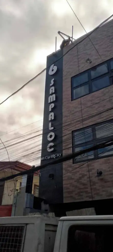 Vertical building signage for 6 SAMPALOC, bold white letters, wires visible, cloudy sky in Ortigas by M&G Global Ads.