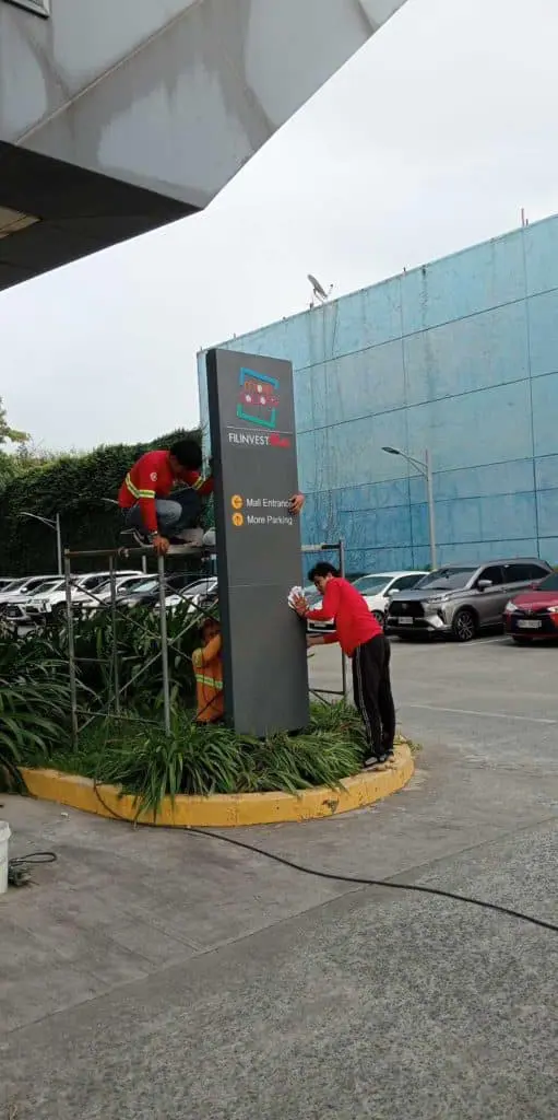Filinvest pylon signage maintenance at mall entrance in Ortigas by M&G Global Ads, with workers on scaffold and cars in background.