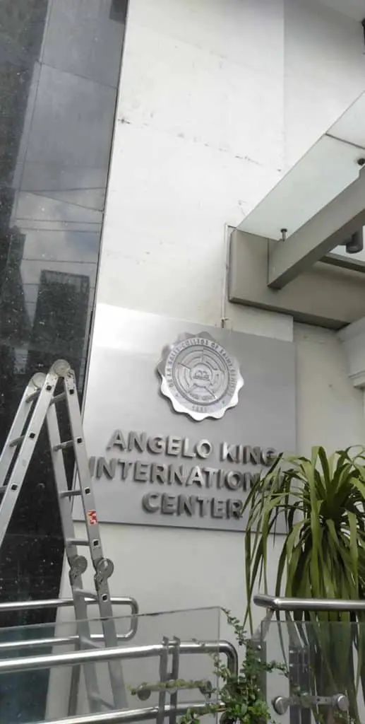 Metallic building signage for ANGELO KING INTERNATIONAL CENTER in Makati on a white wall with ladder and potted plant by M&G Global Ads.