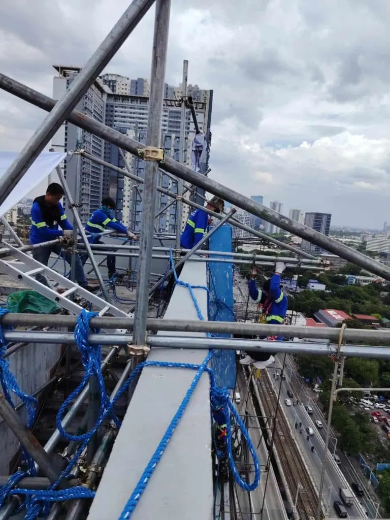 Construction workers install acrylic signage for client on scaffolding in Makati, cityscape below on cloudy day by M&G Global Ads.