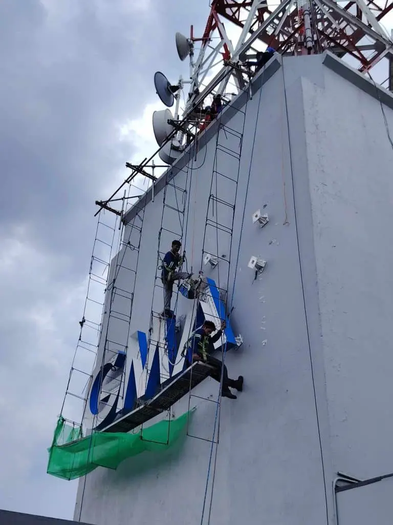 Workers installing blue metallic outdoor business signage on tall building with antennas in Ortigas, by M&G Global Ads.