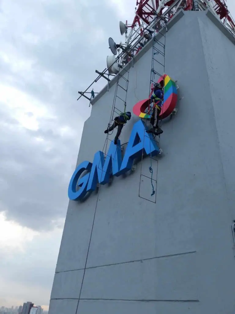 GMA metal building signage installation in Quezon City by M&G Global Ads, workers on harnesses against cloudy sky.