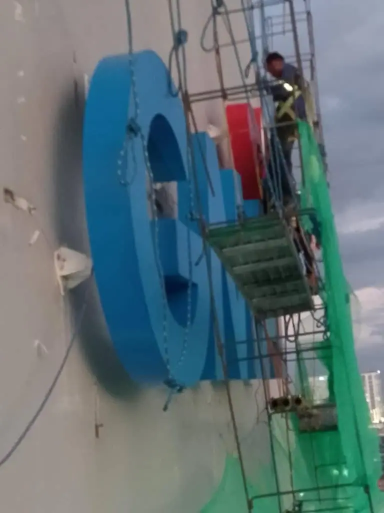 Worker installs large blue and red 3D acrylic signage on building in Ortigas, with ropes and netting by M&G Global Ads.