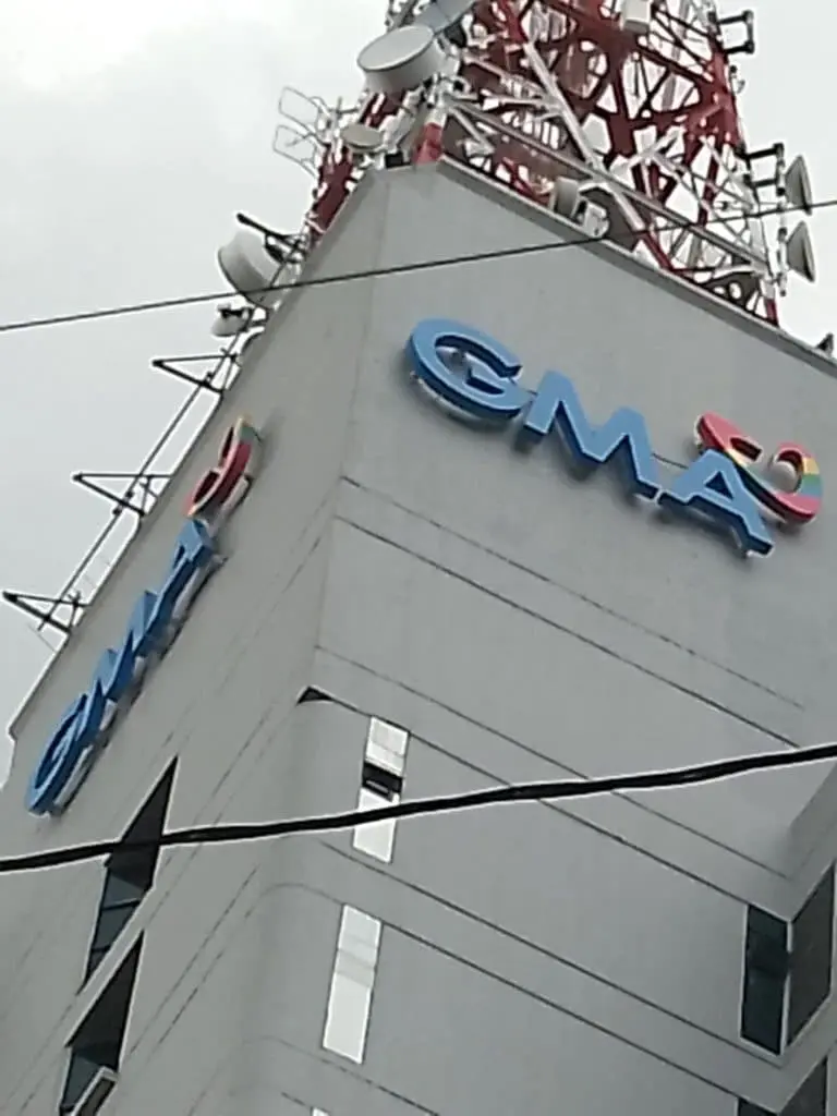 GMA acrylic signage on tall gray building with heart logo, satellite dishes, Quezon City skyline view by M&G Global Ads.