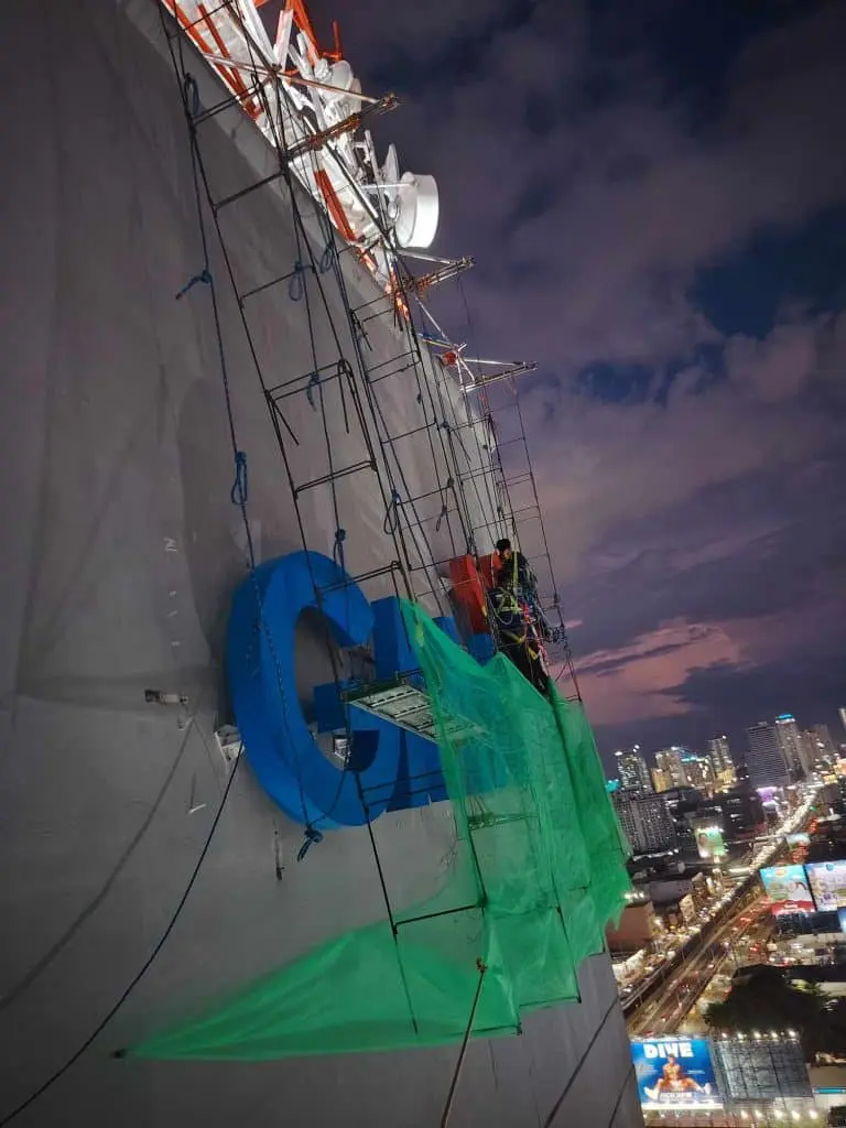 Workers install large blue LED building signage at night in Ortigas with green safety nets, city view—by M&G Global Ads.