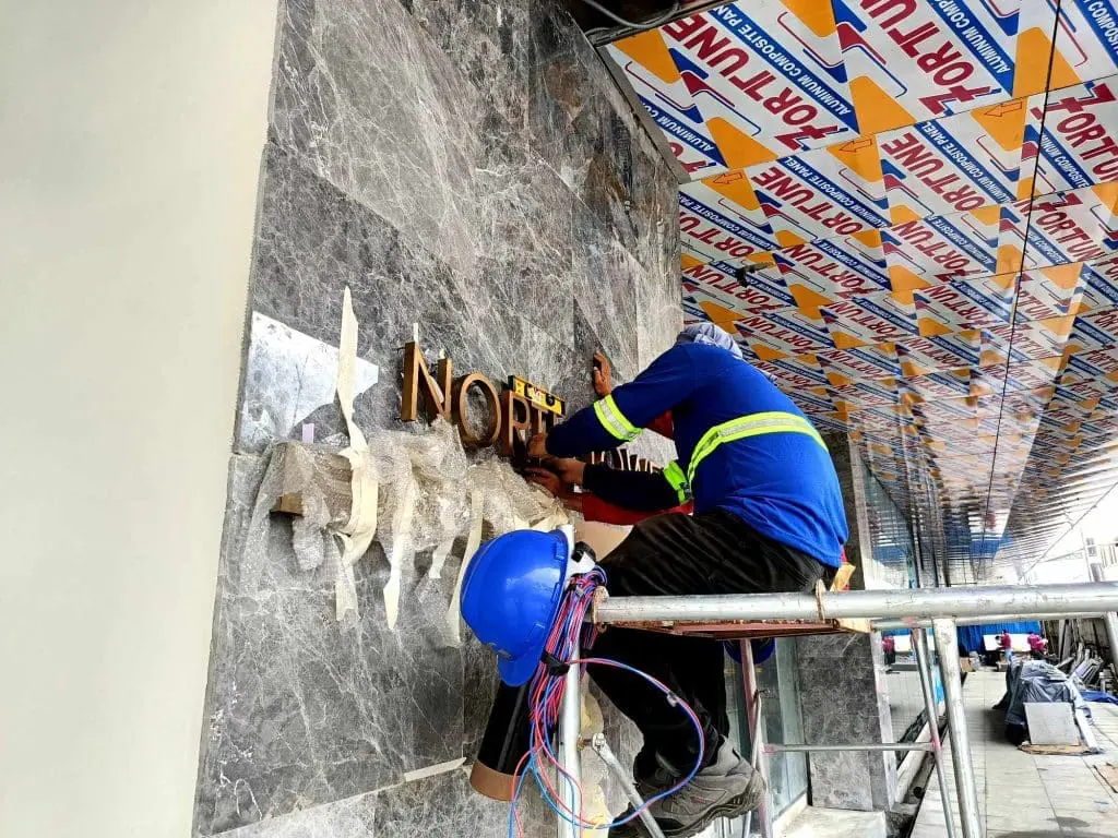 Metallic building signage installation spelling NORTH on marble wall in Makati; worker in blue uniform on scaffolding by M&G Global Ads.