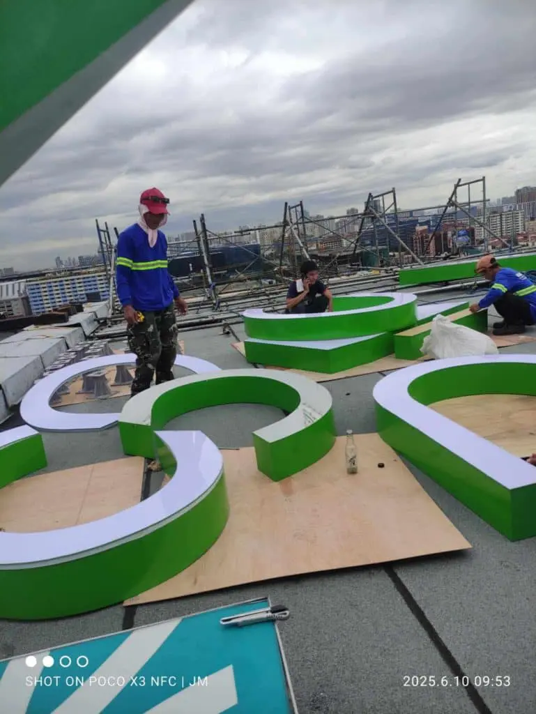 Workers installing green and white acrylic building signage on rooftop in Makati, with city backdrop. By M&G Global Ads.