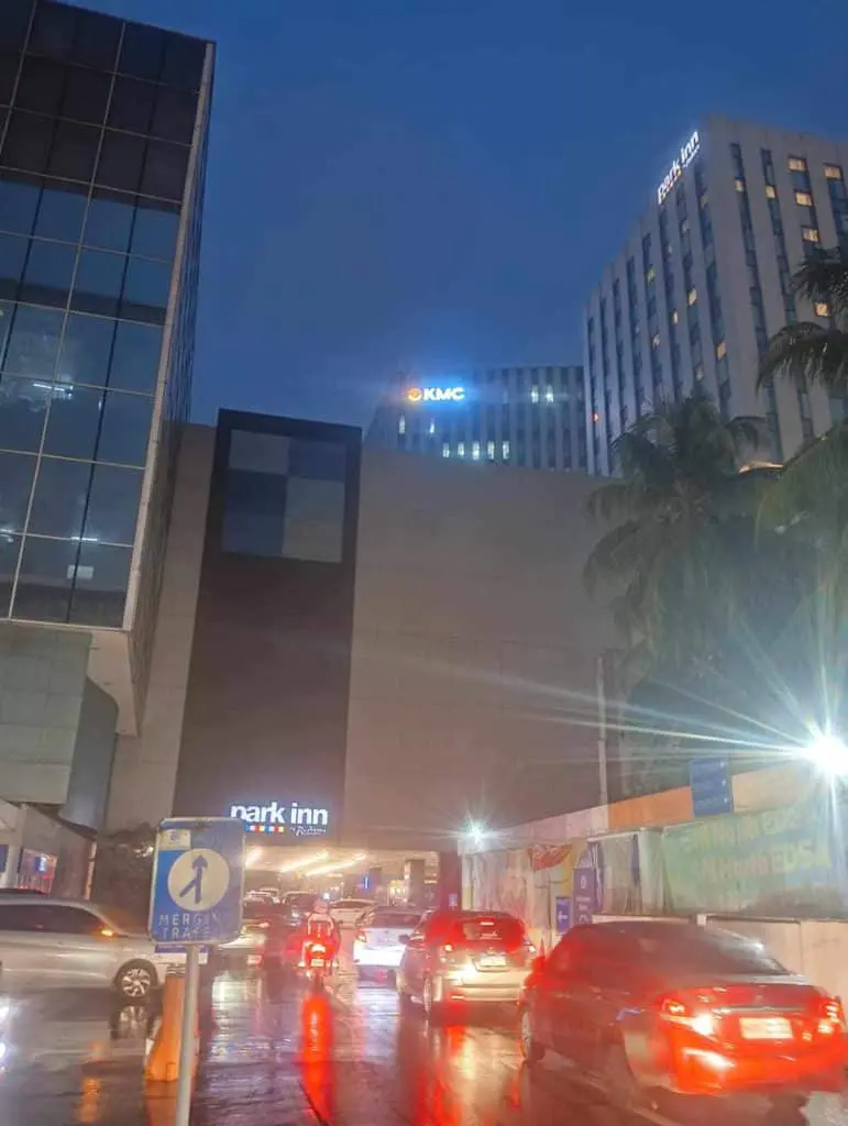 Cars on a wet Makati road with LED signage for KMC and Park Inn on buildings, palm trees visible. by M&G Global Ads.