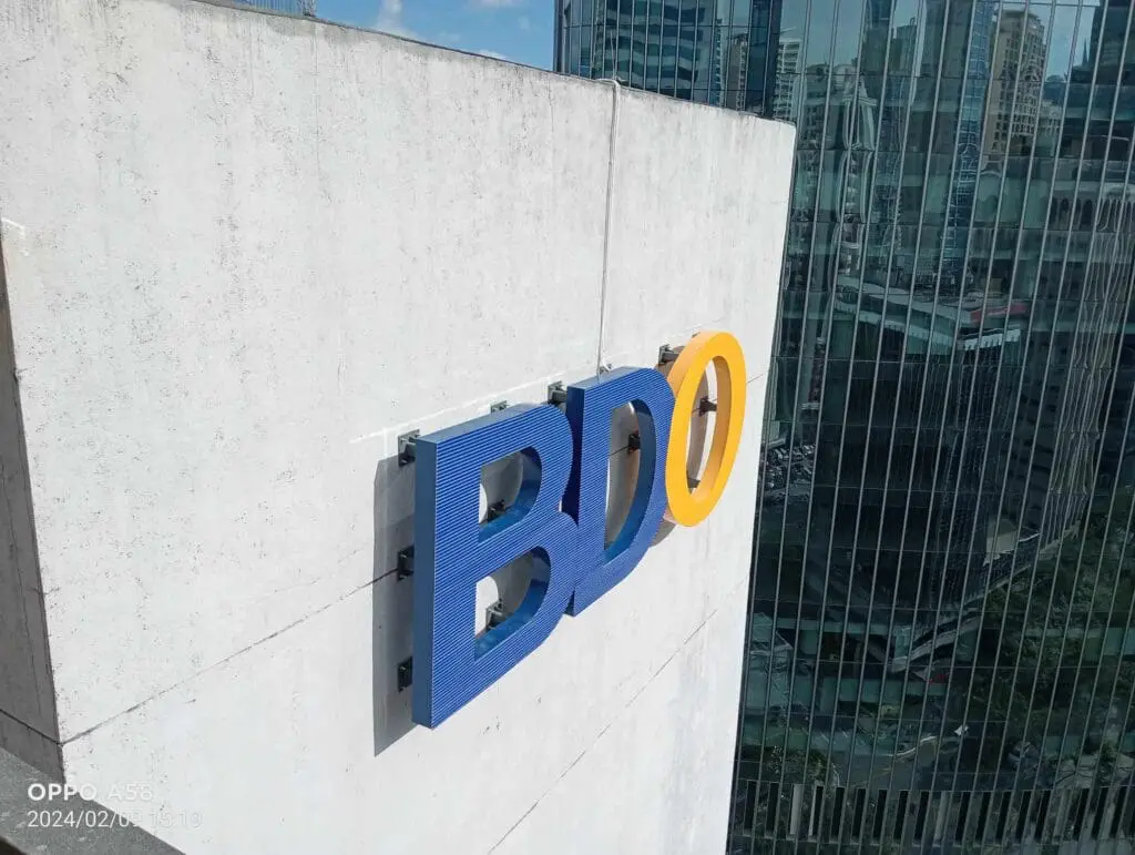 BDO outdoor signage on concrete wall in Makati with glass office buildings, sky reflections—by M&G Global Ads.
