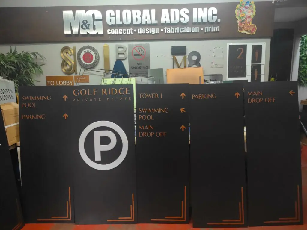 Acrylic and stainless steel wayfinding directional signboards for Golf Ridge Private Estate are showcased in front of the M&G Global Ads Inc. office counter, with multiple signage samples on display behind the counter. Location: Metro Manila, sign maker Philippines by M&G Global Ads.