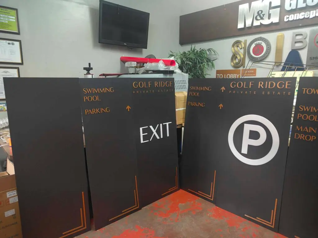 Indoor acrylic wayfinding directional signage for Golf Ridge Private Estate, featuring black and orange “Swimming Pool Parking,” “Exit,” and large “P” parking signs, propped up near a wall and boxes in the Philippines; sign maker Philippines by M&G Global Ads.