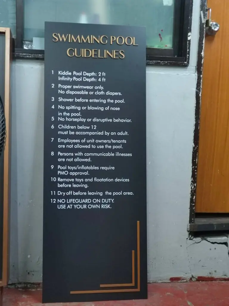 Indoor safety signage, black acrylic Swimming Pool Guidelines sign listing 12 pool rules in white text including swimwear requirements, no spitting, age limits, and pool use at your own risk, propped against a wall near a wooden door in Metro Manila. Sign maker Philippines by M&G Global Ads.