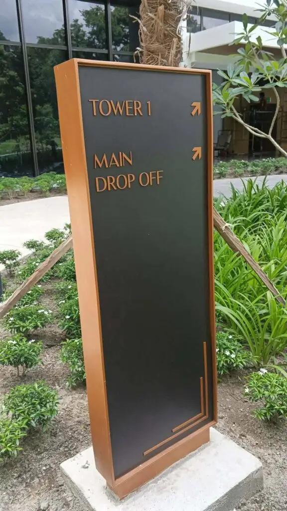 Outdoor wayfinding pylon sign with black and bronze finish, featuring directional arrows pointing right and labels for TOWER 1 and MAIN DROP OFF. Surrounded by green plants, soil, and a building with glass windows in the background in Metro Manila. Sign maker Philippines by M&G Global Ads.