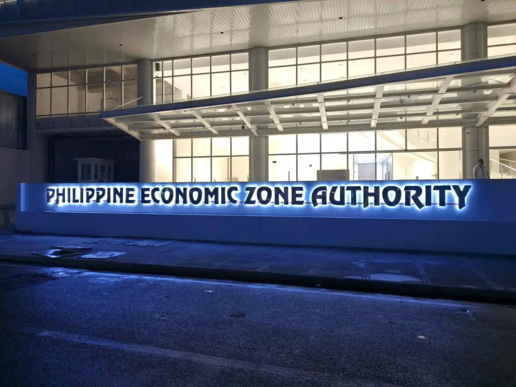 PHILIPPINE ECONOMIC ZONE AUTHORITY acrylic LED building signage in blue and white letters brightly illuminated at night on a modern facade, interior visible through large glass windows; location: Philippines; sign maker Philippines by M&G Global Ads.