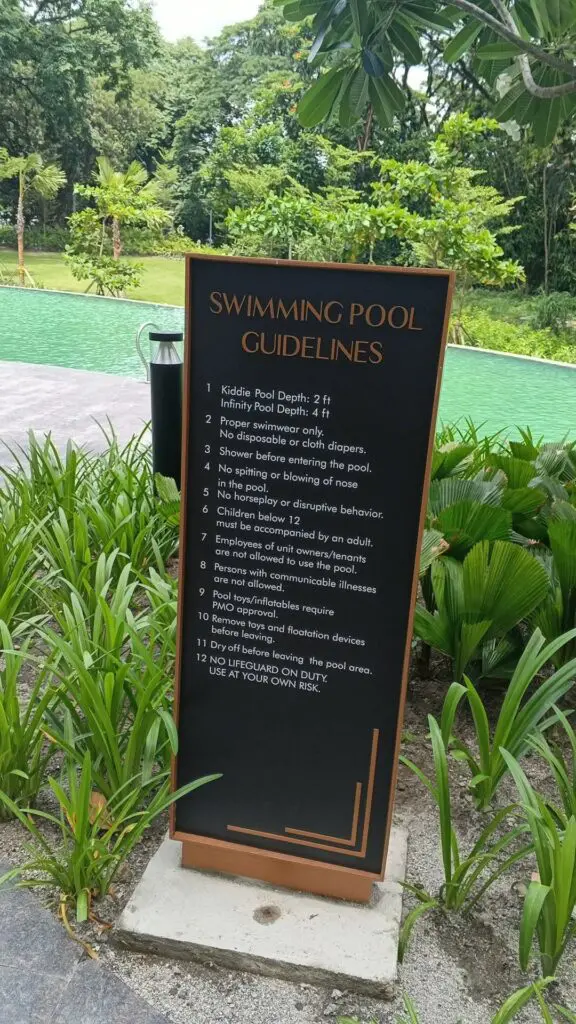 Outdoor safety signage by a swimming pool in Metro Manila, featuring pool rules and guidelines such as pool depths, proper attire, supervision requirements, and prohibitions on food, pets, and running; signage is likely made of durable acrylic or aluminum material with clear printed instructions, surrounded by greenery and a visible swimming pool backdrop; sign maker Philippines by M&G Global Ads.