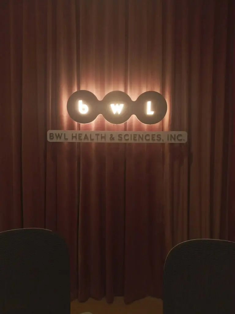 LED acrylic indoor signage for BWL HEALTH & SCIENCES, INC., featuring a glowing "bwl" sign with illuminated dots above an acrylic panel stating the company name, all mounted against a brown curtain background in Metro Manila. Sign maker Philippines by M&G Global Ads.