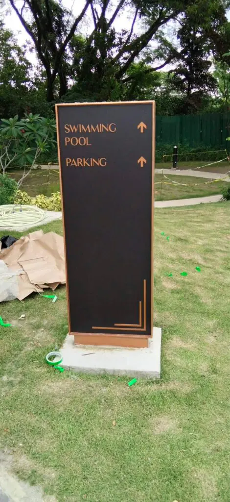 Outdoor wayfinding sign, black and gold, possibly brass or aluminum, with arrows pointing up to indicate directions to the swimming pool and parking area; installed on grass with landscaped greenery and trees in the background in the Philippines. Sign maker Philippines by M&G Global Ads.
