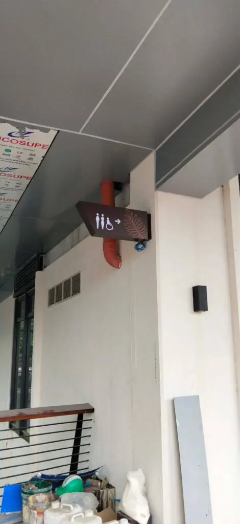 Acrylic indoor restroom wayfinding sign with brown background and white icons for men's, women's, and accessible restrooms, plus a plus sign. The sign is mounted under a gray ceiling near a wall with exposed pipes and cleaning supplies visible below. Location: Philippines. Sign maker Philippines by M&G Global Ads.