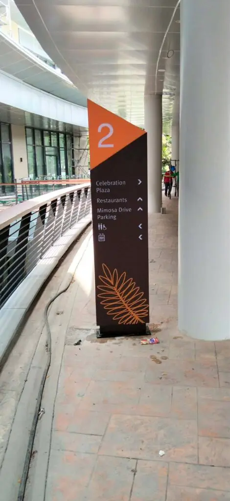 Outdoor wayfinding acrylic sign with an orange top and the number 2, featuring white icons and text for directions to Celebration Plaza, restaurants, Mimosas Drive, and parking. Decorative leaf outline at the base. Sign maker Philippines. Metro Manila. By M&G Global Ads.