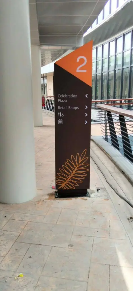 Wayfinding outdoor pylon signage with an orange acrylic top marked "2" directing to Celebration Plaza and Retail Shops, featuring a leaf design at the base, installed on a paved walkway beside a glass-walled building in Metro Manila, sign maker Philippines by M&G Global Ads.
