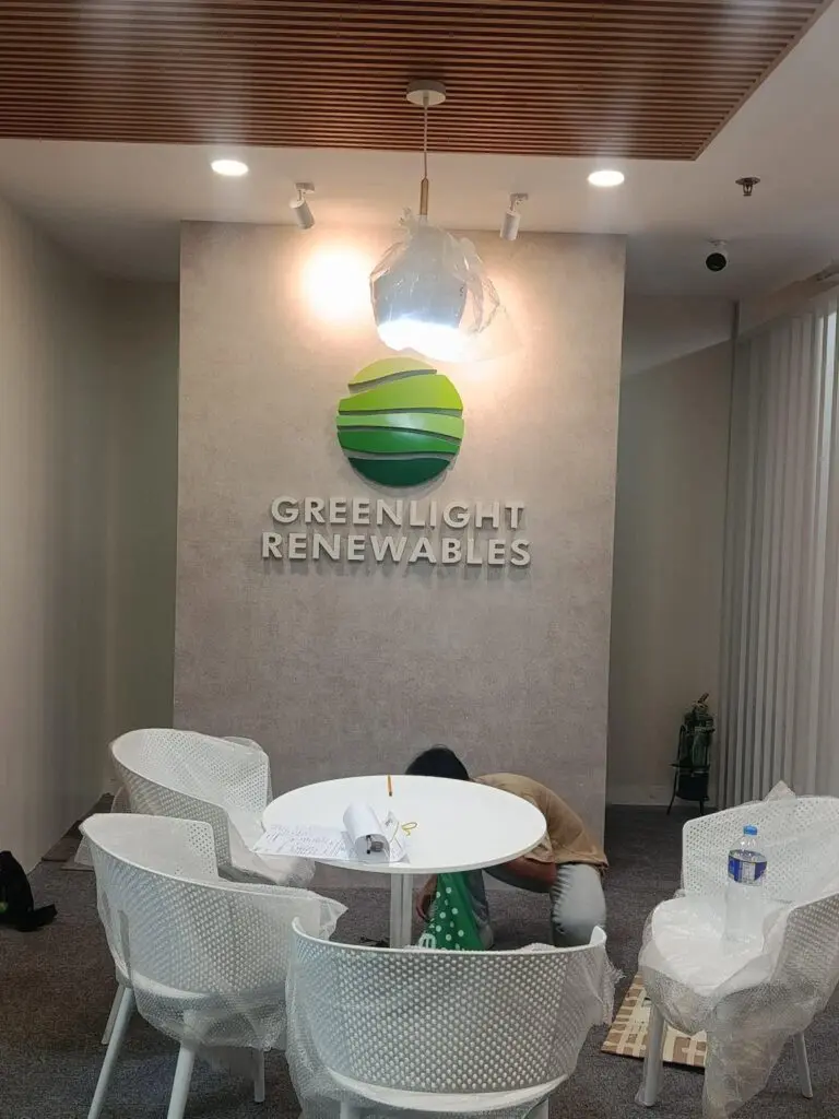 A person crouches near a round table in an office lobby with “Greenlight Renewables” acrylic indoor signage and a green logo above on the wall. White chairs and paperwork are on the table, with office lights and ceiling panels visible. Sign maker Philippines by M&G Global Ads.
