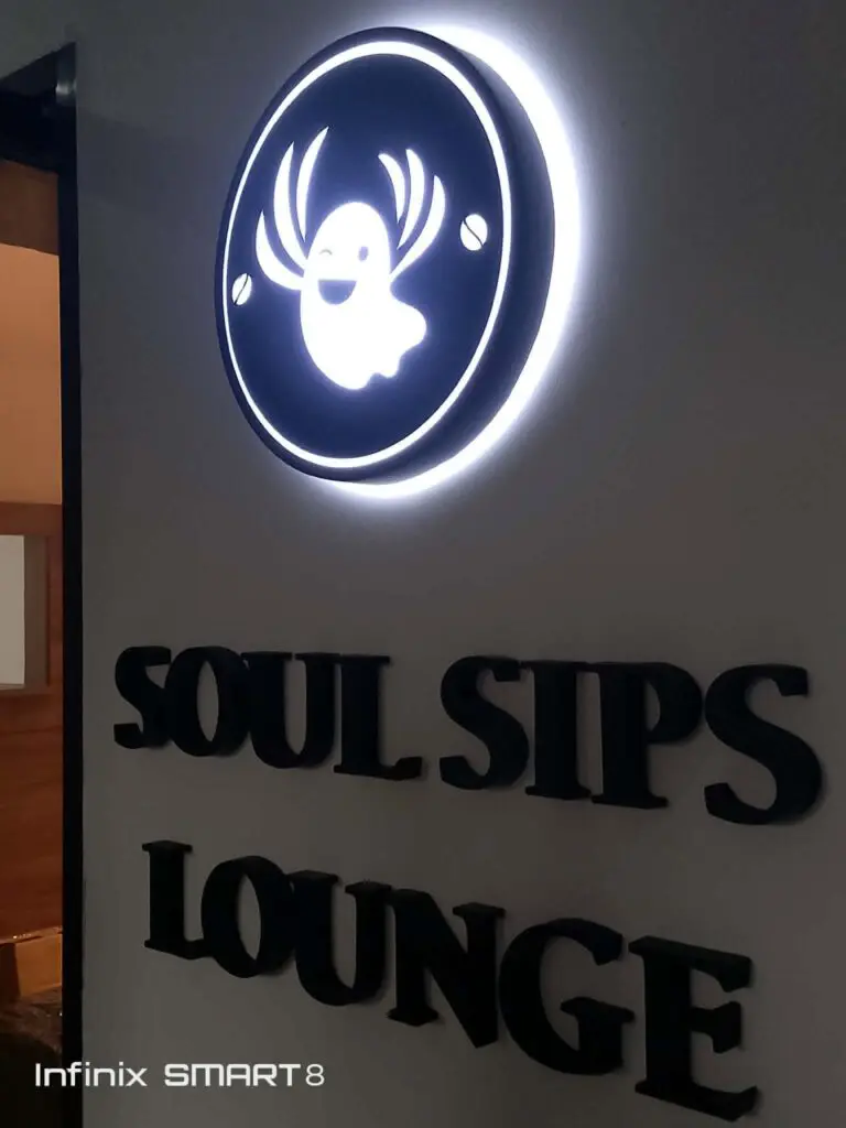 LED acrylic signage for SOUL SIPS LOUNGE featuring a glowing smiling white ghost with antlers above illuminated text, mounted on an indoor wall with part of an open door visible; Infinix SMART8 watermark present. Sign maker Philippines by M&G Global Ads. Location: Metro Manila.