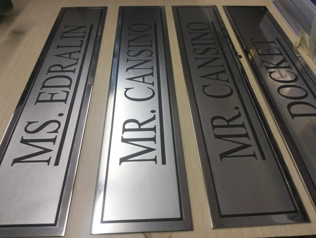 Stainless steel engraved nameplates signage on a wooden surface displaying the names MS. EDRAILN, MR. CANSINO, MR. CANSINO, and MR. DOCKET in black capital letters; office door or room wayfinding signage by sign maker Philippines in Metro Manila by M&G Global Ads.