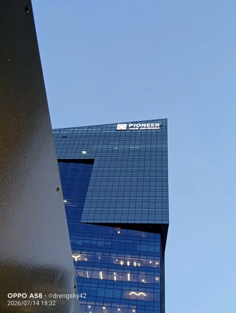 Pioneer Your Insurance acrylic building signage on a modern glass building exterior against a clear blue sky in Metro Manila, viewed from a low angle with part of a metallic structure in the left foreground, sign maker Philippines by M&G Global Ads.