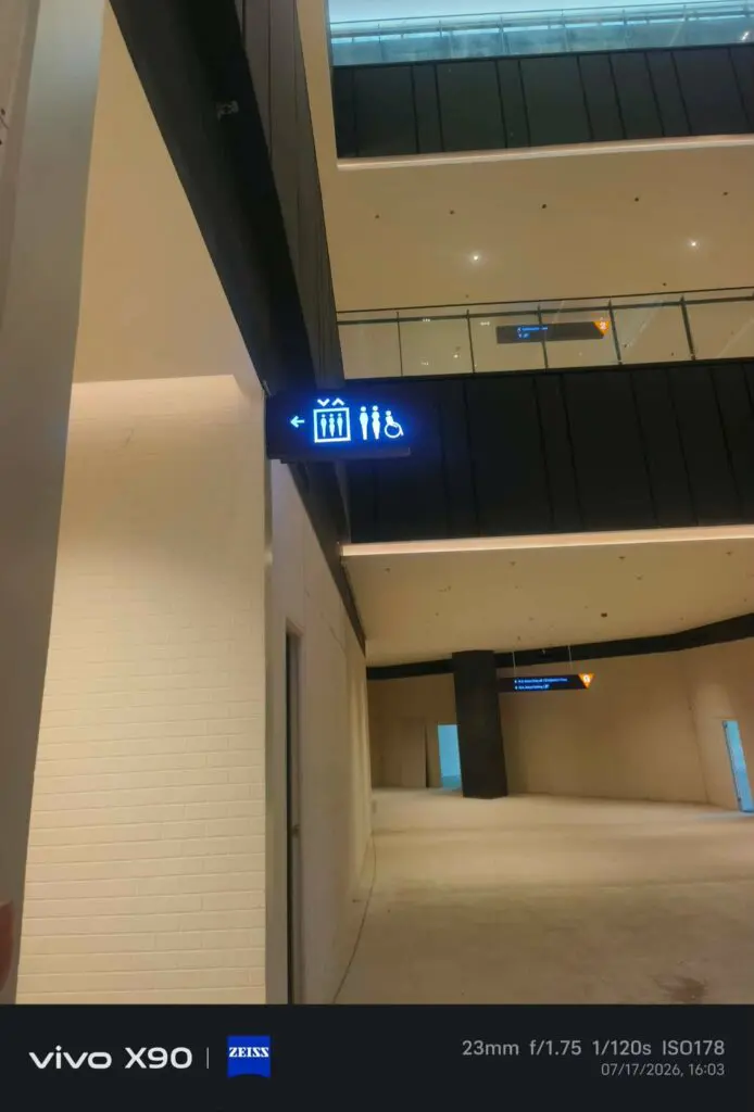 A hallway in a modern building in Metro Manila features indoor wayfinding acrylic signage with icons for a gift shop, restrooms, and accessible facilities. The photo details and timestamp are visible at the bottom. Sign maker Philippines by M&G Global Ads.