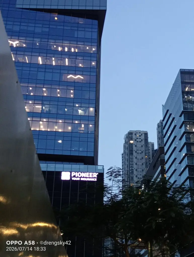 Cityscape at dusk in Metro Manila featuring tall glass buildings, including one displaying a lit LED building sign with the text "PIONEER YOUR EXPERIENCE." Trees and a reflective surface are visible in the foreground under a clear blue sky. Sign maker Philippines by M&G Global Ads.