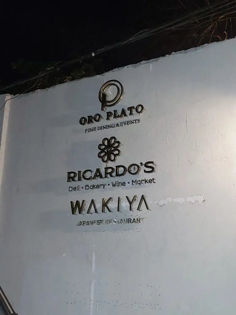 Indoor building signage with gold lettering for Oro Plato Fine Dining & Events, Ricardos Deli-Bakery-Wine-Market, and Wakiya Japanese Restaurant on a white wall, created by sign maker Philippines, Metro Manila location. Signage material features metallic or brass finish. By M&G Global Ads.