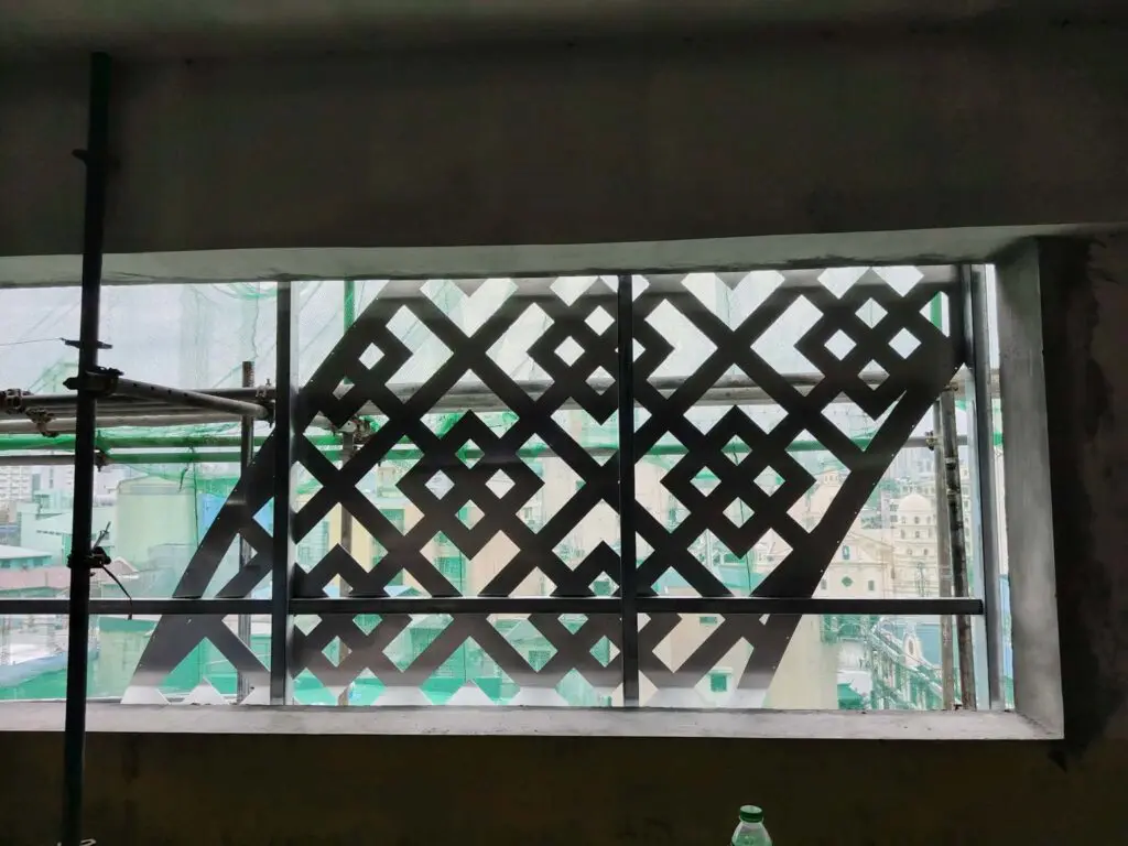 Indoor building signage with a decorative geometric metal grille window, letting in natural light, with a view of buildings and green netting outside indicating construction work in Metro Manila. A plastic bottle is placed on the windowsill inside. Sign maker Philippines by M&G Global Ads.