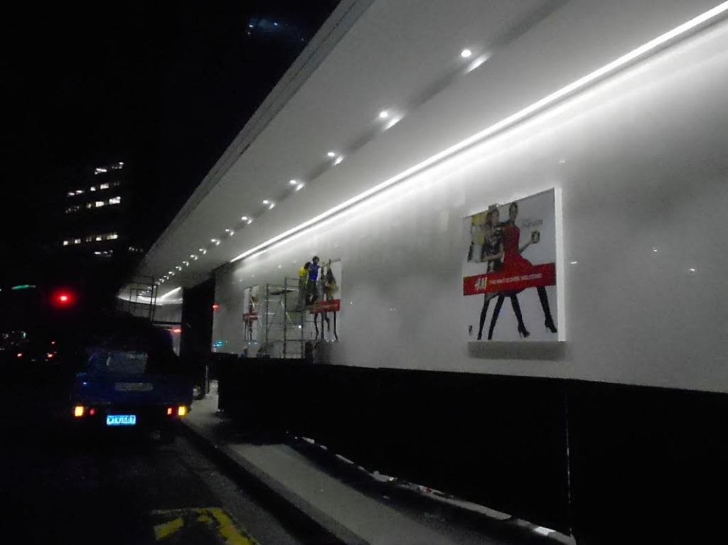Workers install illuminated building signage at night in Makati with a blue car nearby, by M&G Global Ads.