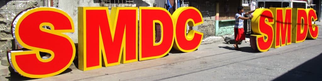 Acrylic SMDC and SMDS signage in bold red and yellow on a sunny Quezon City street, with passerby and dog—by M&G Global Ads.