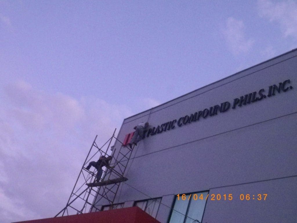 Metallic signage installation for PLASTIC COMPOUND PHILS, INC. in Quezon City by M&G Global Ads; purple sky, workers on scaffolding.