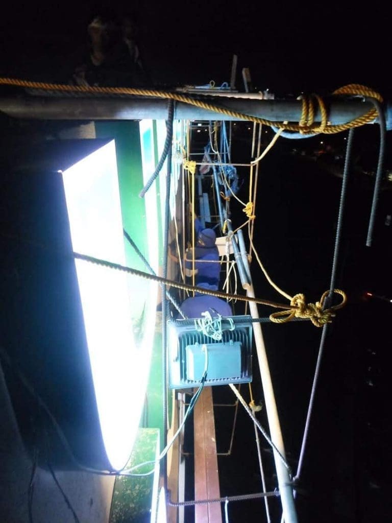 Acrylic signage repair at night with tools and ropes, worker’s legs visible, Ortigas city view by M&G Global Ads.