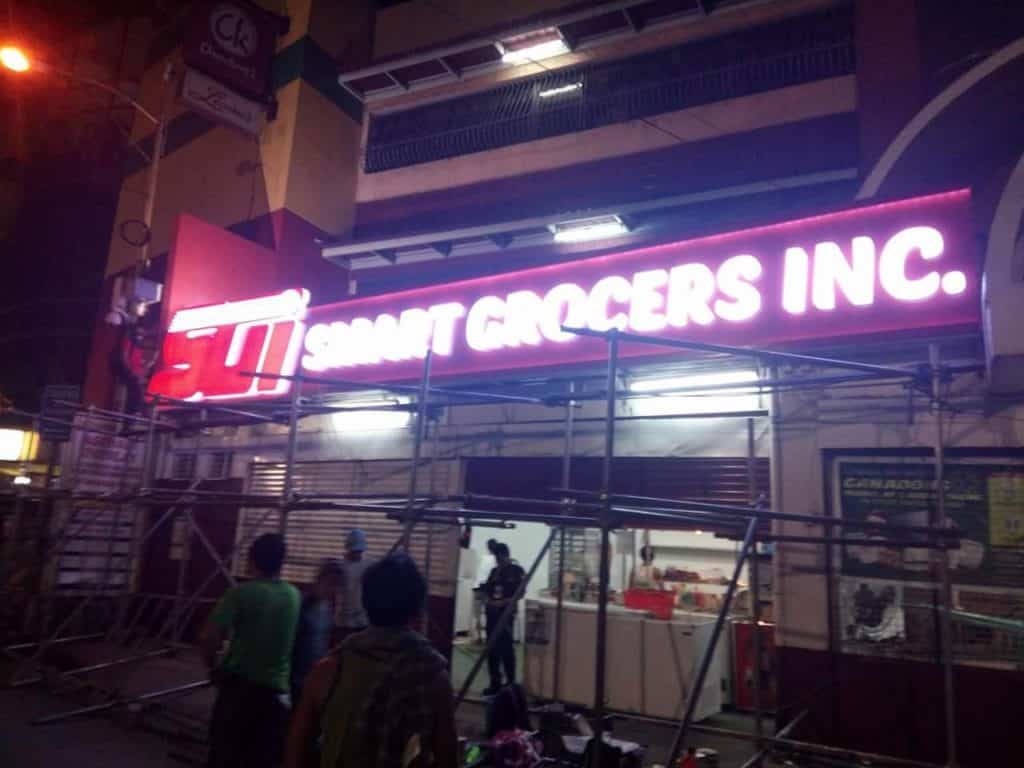 Smart Grocers Inc. LED outdoor signage at night with people near scaffolding in Mandaluyong, by M&G Global Ads.
