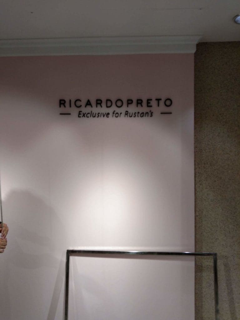 Black metallic indoor signage for Rustan’s on a pink wall in Makati, above a metal clothing rack. by M&G Global Ads.